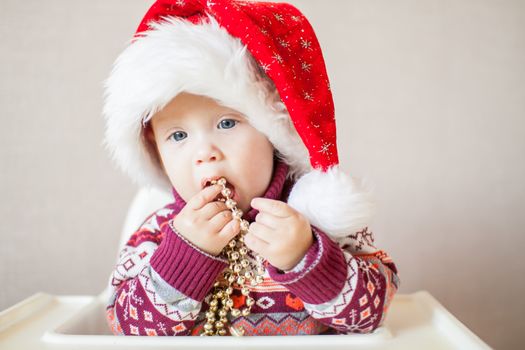 A little baby girl in a New Year's hat of Santa Claus examines and plays with New Year's decorations. Merry Christmas and Happy New Year greetings.