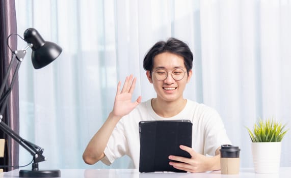 Happy Asian young business handsome man work from home office wear glasses, t-shirt comfortable he smiling and using a black modern smart digital tablet computer raise hand to say hello team on desk