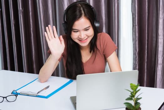 Work from home, Smiling Happy Asian business young beautiful woman sitting on desk workspace wearing headphones video call conference with laptop computer raise hand saying hi team at home office