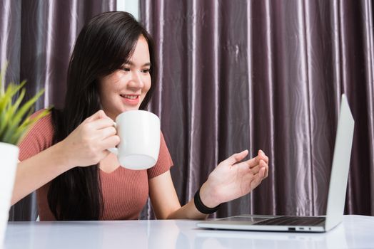 Work from home, Smiling Happy Asian business young beautiful woman sit on desk workspace video call conferencing by webcam laptop computer technology and raise your hand explain the job at home office