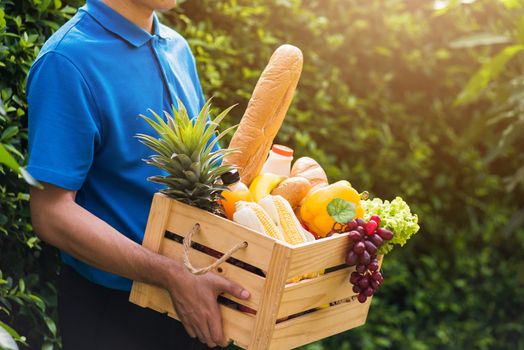 Asian man farmer wears delivery uniform he holding full fresh vegetables and fruits in crate wood box in hands ready give to customer harvest organic food on the garden place green leaves background