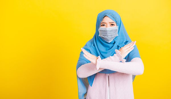 Asian Muslim Arab, Portrait of happy beautiful young woman Islam religious wear veil hijab and face mask protect she quarantines disease coronavirus show hand say no sign, studio shot isolated yellow