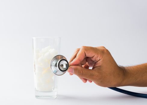 Hand of doctor hold stethoscope check on glass full of white sugar cube sweet food ingredient, isolated on white background, health high blood risk of diabetes and calorie intake unhealthy drink