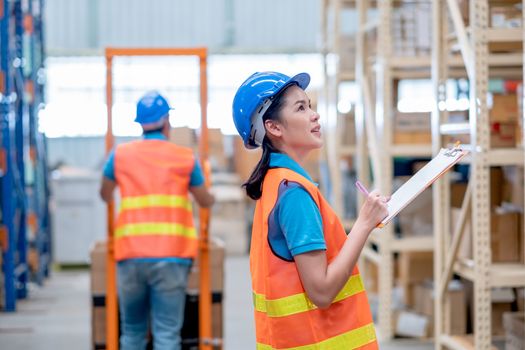 Warehouse woman or factory worker with blue hard hat and uniform stand in front co-worker with cart to check package in workplace. Concept of good management and happiness in  industrial business.