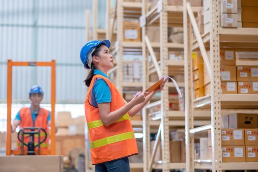 Warehouse woman or factory worker with blue hard hat and uniform stand in front co-worker with cart to check package in workplace. Concept of good management and happiness in  industrial business.