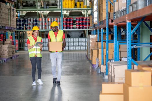 African American warehouse man and woman or factory worker work and discuss together about product box that man holding and walk together in workplace. Concept of good management of staff working.
