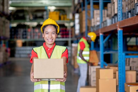 African American warehouse worker or factory woman hold box and action of give to camera in front man to delivery with happiness. Concept of good management of staff work in industrial business.