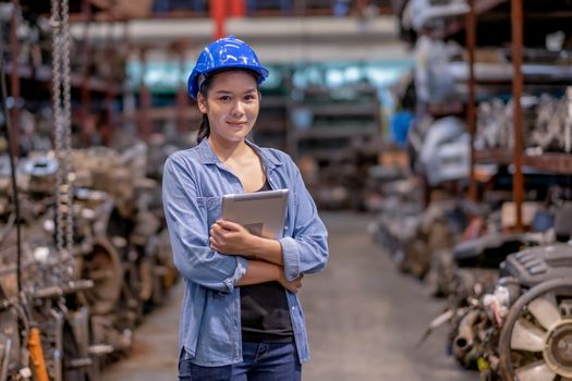 Portrait of factory worker woman with blue hardhat holds tablet and stand in automotive parts workplace area and look to camera. Concept of confident of working with warehouse industrial business.
