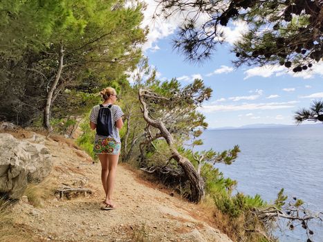 Young active feamle tourist wearing small backpack walking on coastal path among pine trees looking for remote cove to swim alone in peace on seaside in Croatia. Travel and adventure concept.
