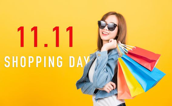 Asian portrait happy beautiful young woman smiling stand with sunglasses excited holding shopping bags multi color with 11.11 shopping day text on space, studio shot isolated yellow background