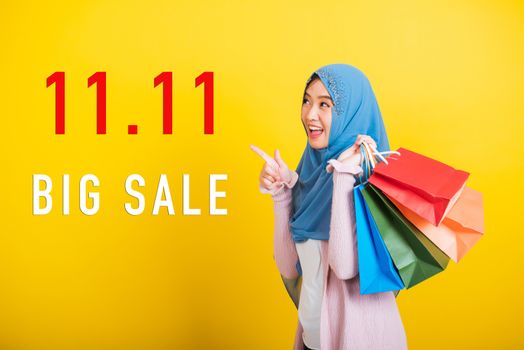 Asian Muslim Arab, Portrait of happy beautiful young woman Islam religious wear veil hijab funny smile she holding colorful shopping bags with 11.11 big sale text at side isolated on yellow background