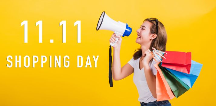 Portrait Asian beautiful happy young woman smiling hold shopping bags multi-color and shouting in megaphone with 11.11 shopping day text at side isolated on yellow background