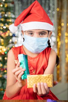 Girl kid in medical mask with Christmas hat and decorated background opening gift box - concept of Gift sharing, distant Xmas celebration due to ccoronavirus or covid-19 pandemic