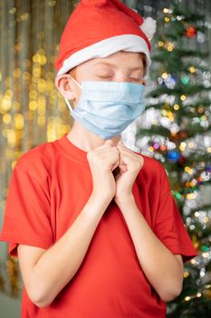 Kid in medical mask and Santa hat praying for santa on Christmas decorated background for gifts