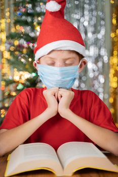Kid with red Santa hat praying in front of Bible on Christmas decorated background with gifts on table - Concept of prayer for a Christmas eve