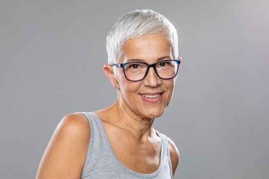Beautiful cute smiling senior woman with short white hair and glasses posing in front of gray background