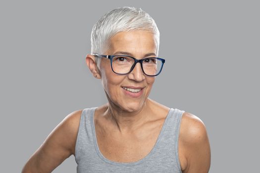 Beautiful smiling cute senior woman with short white hair and glasses posing in front of gray background