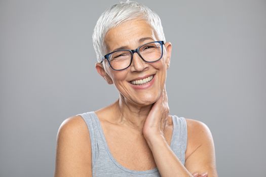 Beautiful cute smiling senior woman with short white hair and glasses posing in front of gray background