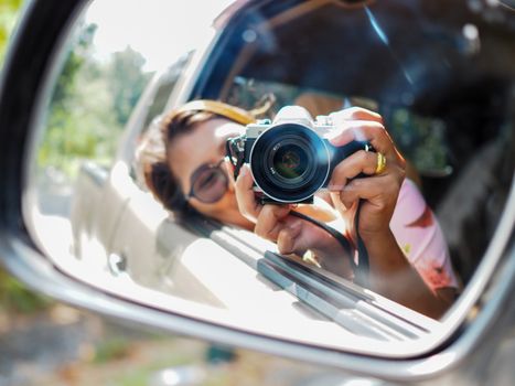 A young woman held a digital camera and took a picture of herself smiling reflected in the car mirror. Focus on camera.