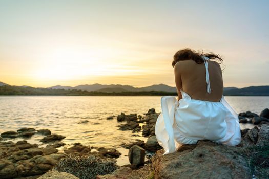 Unrecognizable pensive young caucasian woman in white wedding dress sits on the rocks by the sea at sunset with her head turned down in sadness - Concept of lost love and loneliness