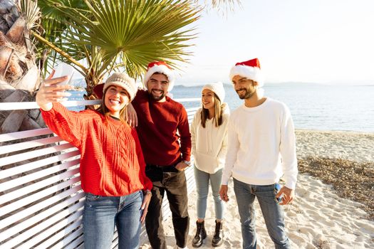 Group of young caucasian friends with Santa Claus hat having fun taking a selfie at the beach in winter for the Christmas holidays - Two couple of people using technology to sharing their journey