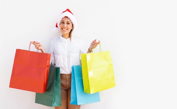 Young beautiful caucasian blonde woman standing on white background with Santa Claus hat holding many colored shopping bag and smiling looking at the camera - Buying Christmas gifts to give happiness