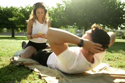 Young caucasian couple trains abdominal muscles in the park - Man doing ABS exercises with his girlfriend keeping time with a smartphone app sitting on his legs - Teamwork is winning work