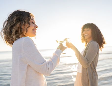Glamour style photograph of two multiracial models, Caucasian and black Hispanic toasting with a glass of white wine by the seawater - Sunset or dawn picture of two women with focus on blonde hair
