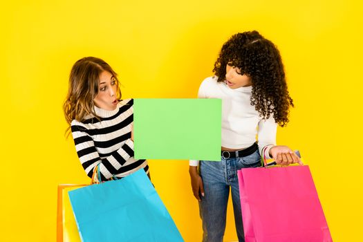 Colors and fun for shopping - Two beautiful young women look at an empty sign that I hold in my hand, ideal as a copy space - Multiracial female models excited and surprised holding some shopping bags