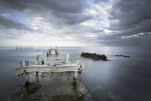 Long exposition of stunning scene of an old abandoned jetty under a dramatic grey cloudy sky with the movement of water sea making silk effect - Focus on the old wooden poles ruined by time