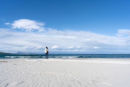 Young unrecognizable woman standing alone by the sea with cloudy sky, looks at the horizon thoughtfully - Pensive single female people in winter on the white beach with Tavolara mountain on background