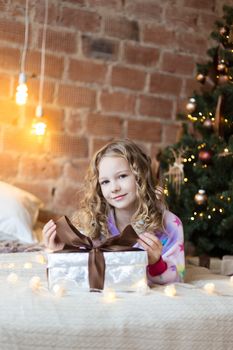 Cute Girl in pajamas lies on bed with a gift box and new year tree and lights behind