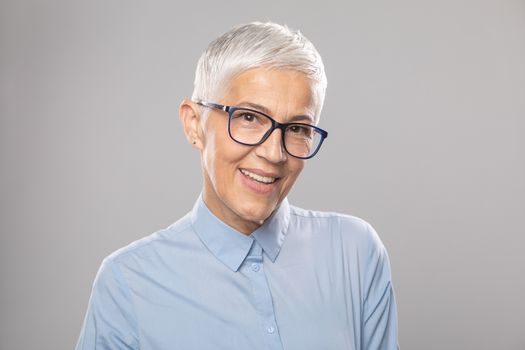 Beautiful smiling cute senior businesswoman with glasses and a blue shirt with short white hair and glasses posing in front of gray background