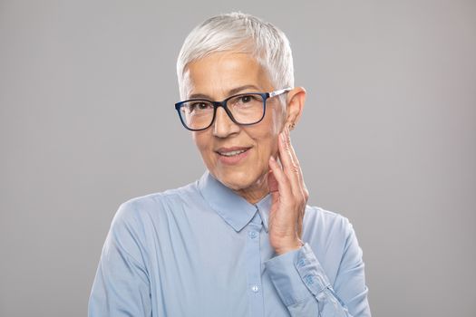 Beautiful smiling cute senior businesswoman with glasses and a blue shirt with short gray white hair and glasses posing in front of gray background