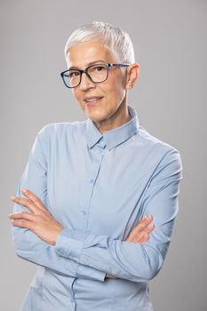 Beautiful smiling cute senior businesswoman with glasses and a blue shirt with short white hair and glasses posing in front of gray background