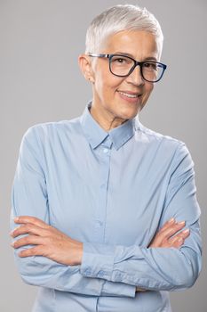 Beautiful smiling cute senior businesswoman with glasses and a blue shirt with short gray white hair and glasses posing in front of gray background