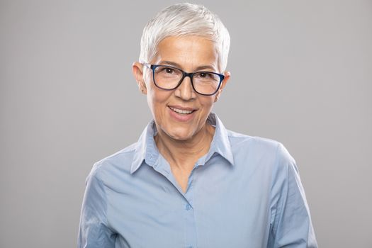 Beautiful smiling cute senior businesswoman with glasses and a blue shirt with short gray white hair and glasses posing in front of gray background