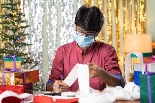 Young man in medical mask busy in calculating holiday expenses bills with christmas decorated background and gifts infront of table