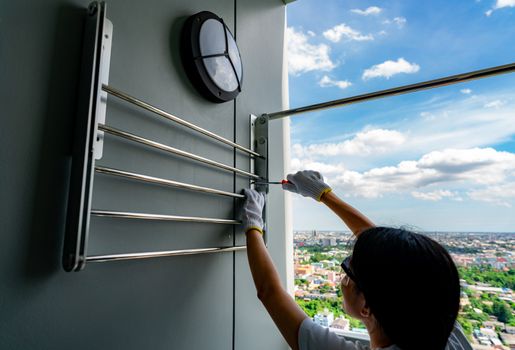 Asian woman use screwdriver screwing screw into the apartment wall for install clothes line. Thai woman wears safety glasses and holding screwdrivers. Woman working home improvement with DIY concept. 