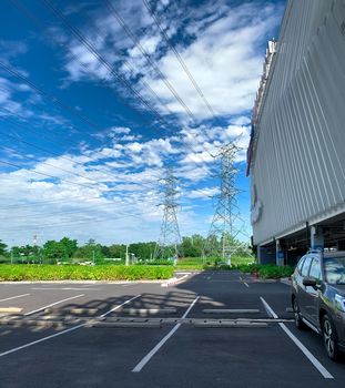 Car parking space at shopping mall for customer service. Outdoors asphalt car parking lot on sunny day with green tree forest and high voltage electric tower. Overhead transmission line. 