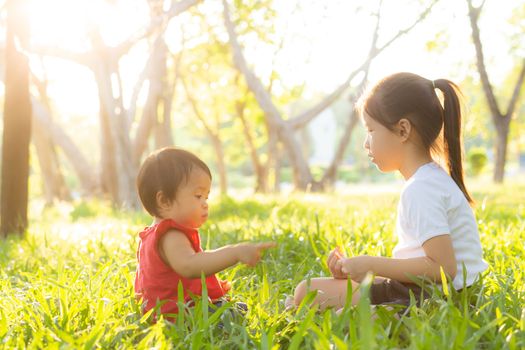 Beautiful young asian kid sitting playing in summer in the park with enjoy and cheerful on green grass, children activity with relax and happiness together on meadow, family and holiday concept.