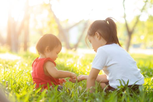 Beautiful young asian kid sitting playing in summer in the park with enjoy and cheerful on green grass, children activity with relax and happiness together on meadow, family and holiday concept.