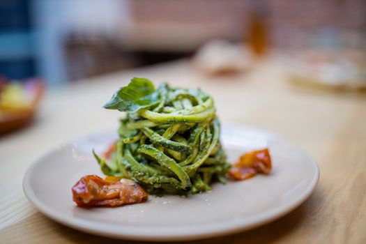 Close up picture of zucchini noodles with a spinach leaf on top and on a wooden restaurant table with blurry tables, chairs, bottles, and a pizza as background