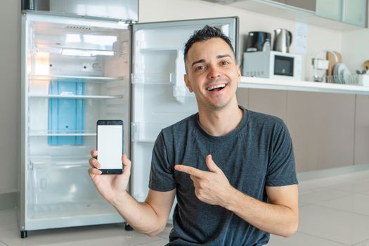 A young guy orders food using a smartphone. Empty refrigerator with no food. Food delivery service advertisement.