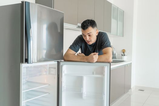 The guy is sad near the empty refrigerator without food. Food delivery service advertisement.