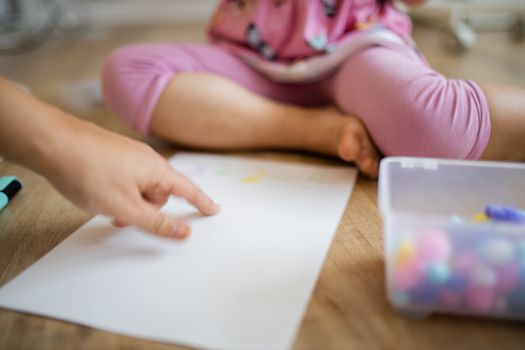 Picture of a hand pointing at a paper sheet on the wooden floor alongside the legs of a little girl and a plastic box full of colorful cotton balls