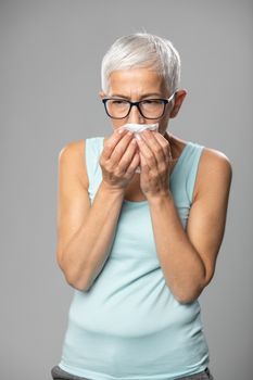 Senior women with short gray hair and glasses coughs and sneezes into a handkerchief, studio shoot, allergies and illness concept