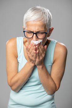 Senior women with short gray hair and glasses coughs and sneezes into a wipe handkerchief, studio shoot, allergies and illness concept