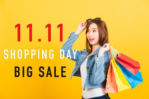 Asian portrait happy beautiful young woman smiling stand with sunglasses excited holding shopping bags multi color with 11.11 shopping day and big sale text on space isolated yellow background