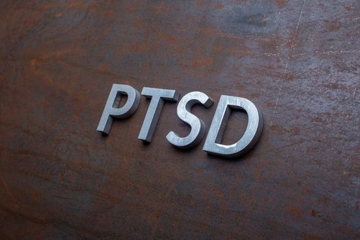 the word PTSD - post traumatic stress disorder - laid with silver metal letters on flat rusted sheet steel background in slanted diagonal perspective.